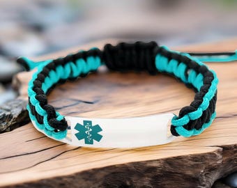 May include: A black and teal paracord bracelet with a white metal plate. The plate features a teal medical symbol. The bracelet is on a wooden surface.