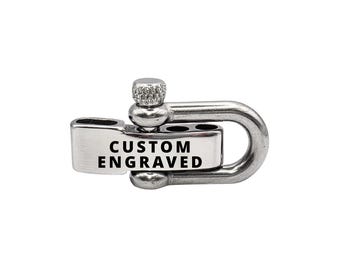 Könnte beinhalten: Ein silberfarbener Schäkelverschluss mit dem schwarzen Text "CUSTOM ENGRAVED". Der Verschluss hat ein abgerundetes, U-förmiges Design mit einem Schraubmechanismus. Der Hintergrund ist weiß.