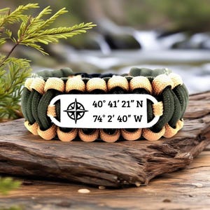 Könnte beinhalten: Ein Survival-Armband mit einem gewebten Design in Olivgrün, Beige und Schwarz. Eine weiße rechteckige Platte in der Mitte zeigt eine Windrose und die Koordinaten 40° 41' 21" N 74° 2' 40" W.