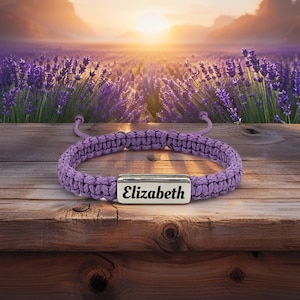 May include: A braided purple bracelet with a silver rectangular plate engraved with the name "Elizabeth". The bracelet is displayed on a wooden surface with a background of a lavender field at sunset.
