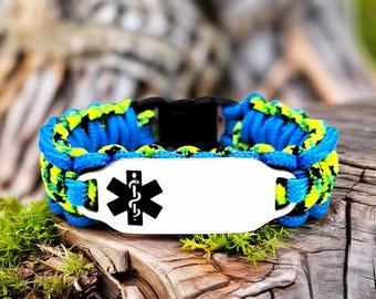 Puede incluir: Una pulsera de paracord azul y amarillo con una placa de metal blanco con un símbolo médico negro. La pulsera tiene un cierre de plástico negro y se muestra sobre una superficie de madera con musgo verde.