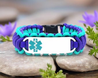 May include: A medical alert bracelet with a white metal plate and a teal medical symbol. The bracelet is made of woven purple and teal paracord and has a black clasp. The bracelet is resting on a stone surface.