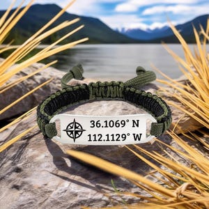 Puede incluir: Una pulsera de paracord verde y negro con una placa plateada. La placa presenta un diseño de rosa de los vientos y las coordenadas 36.1069° N 112.1129° W. La pulsera se muestra sobre una roca con un lago y montañas al fondo.