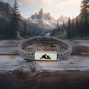May include: Gray braided bracelet with a silver rectangular clasp featuring a black mountain range design. The bracelet is displayed on a rustic wooden surface, with a scenic mountain backdrop.
