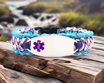 May include: A blue, purple, and white paracord bracelet with a silver medical ID tag. The tag features a purple Star of Life symbol. The bracelet has a black plastic clasp and is displayed on a wooden surface.