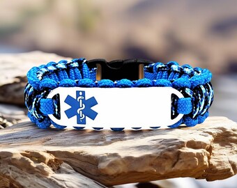 May include: Blue and black paracord bracelet with a white metal plate featuring a blue medical symbol. The bracelet has a black clasp and is displayed on a piece of wood.