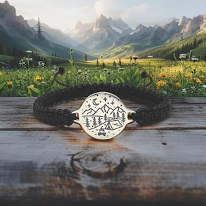 May include: Black braided bracelet with a circular metal pendant featuring a camping scene with mountains, trees, a tent, and stars. The bracelet is set against a wooden surface with a scenic mountain backdrop.