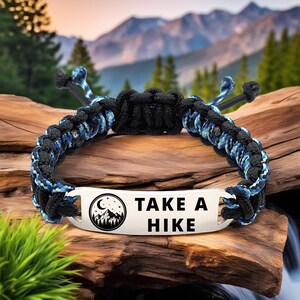 May include: A black and blue braided bracelet with a metal plate that reads "TAKE A HIKE" with a mountain and moon graphic. The bracelet is displayed on a piece of wood with a mountain backdrop.