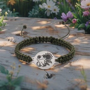 Puede incluir: Una pulsera de macramé verde con un dije plateado con un diseño de rana y girasol. La pulsera se exhibe sobre una superficie de madera con flores en el fondo. La longitud ajustable de la pulsera es de aproximadamente 15 cm.