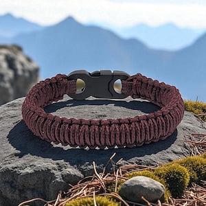 May include: A maroon paracord bracelet with a black plastic clasp. The woven bracelet is resting on a gray rock with a mountain backdrop. The bracelet is a survivalist accessory.