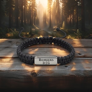 Könnte beinhalten: Ein dunkelgraues geflochtenes Armband mit einer silbernen rechteckigen Platte. Auf der Platte steht der Text "Römer 3,25". Das Armband wird auf einer Holzoberfläche mit einem Waldhintergrund präsentiert.