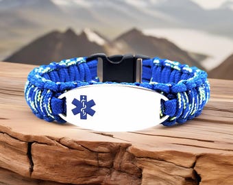 May include: Blue and white paracord medical ID bracelet with a black clasp and a silver plate. The plate features a blue medical symbol. The bracelet is displayed on a wooden surface with a mountain backdrop.