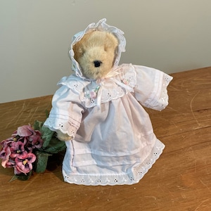 May include: A plush teddy bear wearing a white dress with lace trim and a matching bonnet. The bear has brown eyes and a brown nose. The bear is sitting on a wooden surface.
