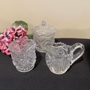 May include: A clear, cut-glass sugar bowl with a lid, a creamer, and a small tumbler. The pieces have intricate starburst and diamond patterns. A bouquet of pink and purple flowers rests to the left of the glassware.
