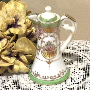 May include: A miniature porcelain pitcher with a lid, featuring a white base, a green rim, and a gold-trimmed handle. The body is decorated with a painted basket of flowers in pink, blue, and green, accented by delicate gold scrollwork patterns.