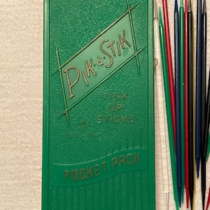 May include: Green plastic case with gold lettering that reads "Pik-a-Stik" and "Pick Up Sticks Pocket Pack". The case contains a set of colorful plastic sticks.