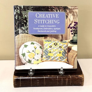 May include: A book titled "Creative Stitching" displayed on a stand. The cover features a wicker chair, two decorative pillows, and the text "A Guide to Cross-stitch, Needlepoint, Embroidery, Appliqué, Patchwork and Quilting."