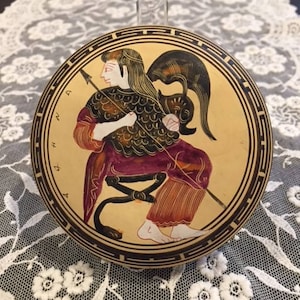 May include: A round ceramic plate with a black and gold border. The plate features a hand-painted image of a Greek goddess holding a spear. The goddess is wearing a purple and gold tunic and a helmet. The plate has the text "ATHENA" written around the border.