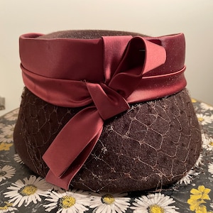 May include: A brown felt hat with a burgundy satin ribbon tied in a bow. The hat has a net-like texture.