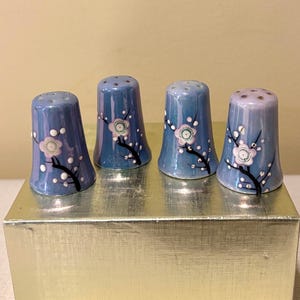 May include: Four blue ceramic thimbles with white and pink floral designs. The thimbles are arranged in a row on a gold surface.
