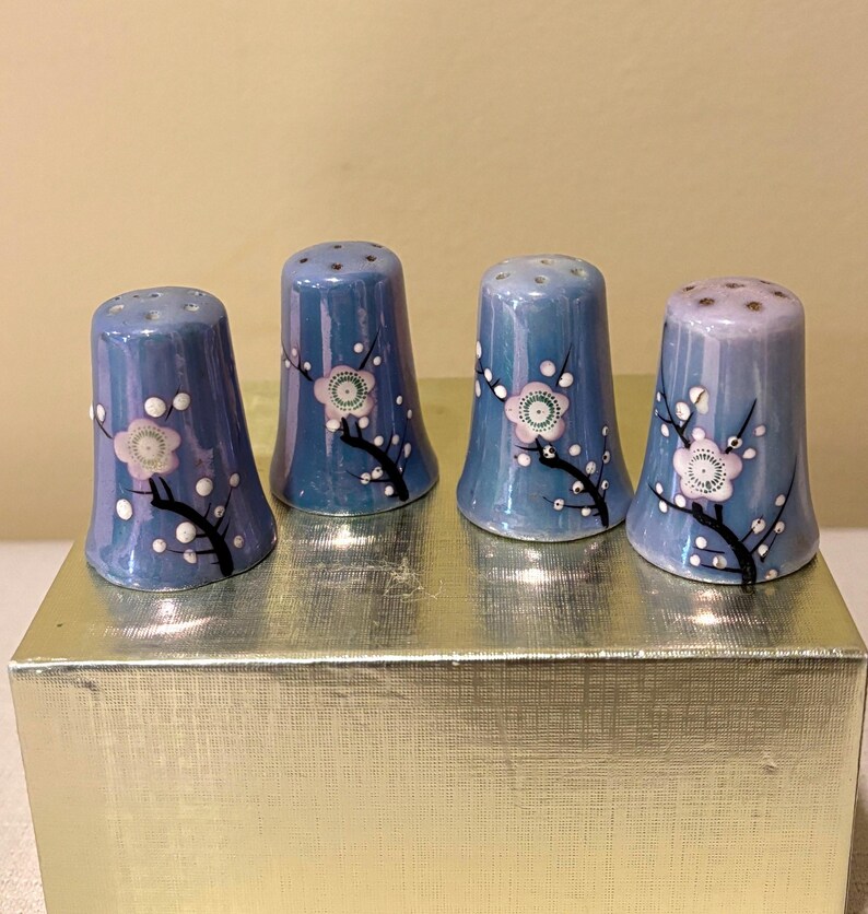 May include: Four blue ceramic thimbles with white and pink floral designs. The thimbles are arranged in a row on a gold surface.