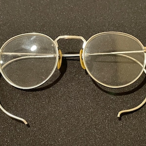 May include: Silver-toned round eyeglasses with thin metal frames and clear lenses. The arms of the glasses are thin and curved.