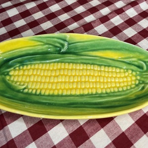 May include: A ceramic serving dish shaped like an ear of corn. The dish is oval, with a yellow corn cob center and green husks. The dish is on a red and white checkered tablecloth.