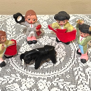 May include: A collection of miniature figurines depicting a bullfight scene. The set includes a black bull and several matador figures in colorful attire, holding capes and other props. The figurines are set on a white lace tablecloth.