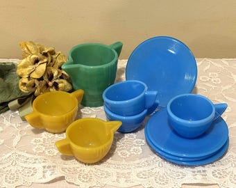 May include: A collection of vintage dishware in shades of blue, yellow, and green. The set includes cups, saucers, and small pitchers. The pieces are arranged on a white lace tablecloth, with dried flowers in the background.