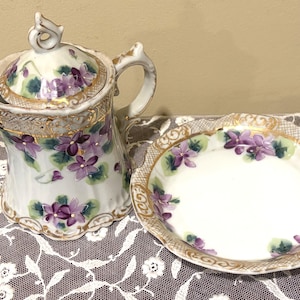 May include: A porcelain creamer pitcher with a matching saucer, both featuring a white base decorated with hand-painted purple violet flowers, green leaves, and ornate gold trim along the scalloped edges and the lid handle.