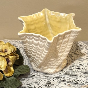 May include: Cream-colored ceramic vase with a textured, shell-like design. The interior is a soft yellow. The vase has a unique, star-shaped top and is likely for decorative purposes.
