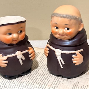 May include: Two ceramic figurines of monks. The figures are wearing brown robes with white rope belts. They have rosy cheeks and are smiling. One figure is a small container with a white rim.