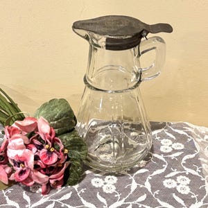 May include: A clear glass pitcher with a metal lid and handle. The pitcher is decorated with vertical lines and sits on a white lace tablecloth. Pink and purple flowers are to the left of the pitcher.