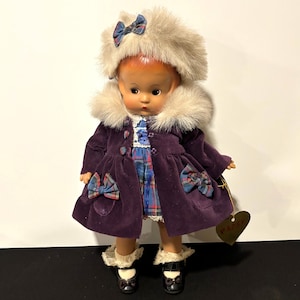 May include: A vintage doll wearing a purple velvet coat with plaid bows and a matching hat with a fur trim. The doll has a plaid dress and black shoes. A heart-shaped tag is attached to the doll.