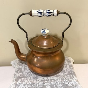May include: A copper tea kettle with a rounded body and a curved spout. The lid features a small white ceramic knob with blue floral accents. The arched metal handle includes a central white ceramic grip decorated with blue leaf patterns.
