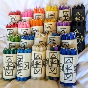 Könnte beinhalten: Ein Set mit 15 kleinen Kerzen, jede in Papier verpackt mit einem anderen Symbol. Die Kerzen sind in verschiedenen Farben, darunter Rot, Rosa, Orange, Gelb, Grün, Blau, Lila, Weiß und Schwarz.