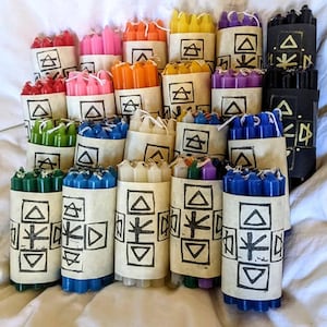 May include: A collection of small, multi-colored taper candles bundled together with paper bands. Each band features a hand-stamped black ink design consisting of geometric shapes, triangles, and a central star-like symbol on a light tan background.