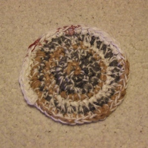 May include: A round crocheted coaster with a brown, white, and gray pattern. The coaster has a slightly raised edge.