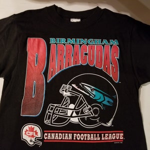 May include: Black t-shirt with a red and blue graphic of a football helmet with a fish inside. The text "BIRMINGHAM BARRACUDAS" is above the helmet. The text "CFL CANADIAN FOOTBALL LEAGUE" is below the helmet.