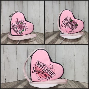 May include: Pink heart-shaped handbag with a black strap and embroidered text that reads "Not Your Sweetheart" and "Dangerous Woman" on the front.