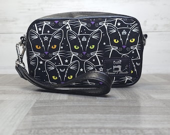 May include: A black rectangular wristlet bag featuring a repeating pattern of cat faces with yellow and orange eyes, white stars, and geometric symbols. A black wrist strap is attached to the side. A label reads Bells Thread & Needle.