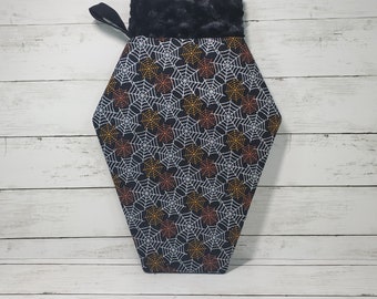 May include: A black and white coffin-shaped stocking with a black fuzzy cuff. The stocking is decorated with a spiderweb pattern in orange and white.