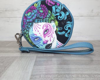 May include: A round zippered pouch with a light blue wrist strap and a silver metal clip. The front features a colorful graphic print of a face with sugar skull makeup, adorned with purple and blue flowers against a black background.