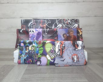 May include: A collection of small, rectangular fabric pouches featuring various horror and pop culture character prints, including scenes with skeletons, ghosts, and movie figures, arranged in a tiered display on a white surface.