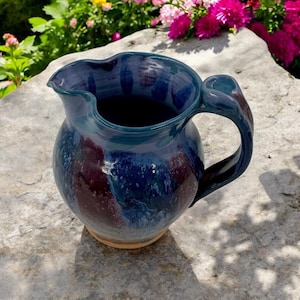May include: A blue and purple ceramic pitcher with a unique, wavy rim and a handle. The pitcher has a speckled glaze and a rustic, handmade look.