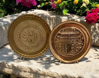 May include: Two decorative brass trays. One tray features intricate Arabic calligraphy in a circular design. The other tray features a raised image of a traditional Korean building with people in front of it.