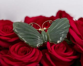 May include: A jade butterfly brooch with gold detailing. The butterfly features carved wings and a detailed body. The brooch is set against a backdrop of red roses, creating a striking contrast. This jewelry piece is a unique accessory.