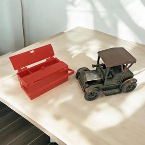 May include: A red metal toolbox with a hinged lid and a handle, and a vintage-style, brown metal toy car.