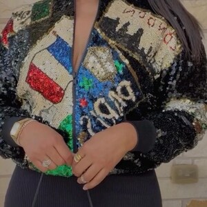 Könnte beinhalten: Eine schwarze Bomberjacke mit Pailletten und Reißverschluss. Die Jacke hat ein farbenfrohes, gesticktes Design mit verschiedenen Flaggen und dem Wort "Ciao". Das Model trägt die Jacke mit einem schwarzen Rock und einer schwarzen Handtasche.