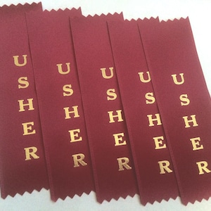 May include: Five burgundy ribbons with gold lettering spelling "USHER". Each ribbon has a zig-zag edge at the top. These ribbons are likely used for event identification or as a decorative element.